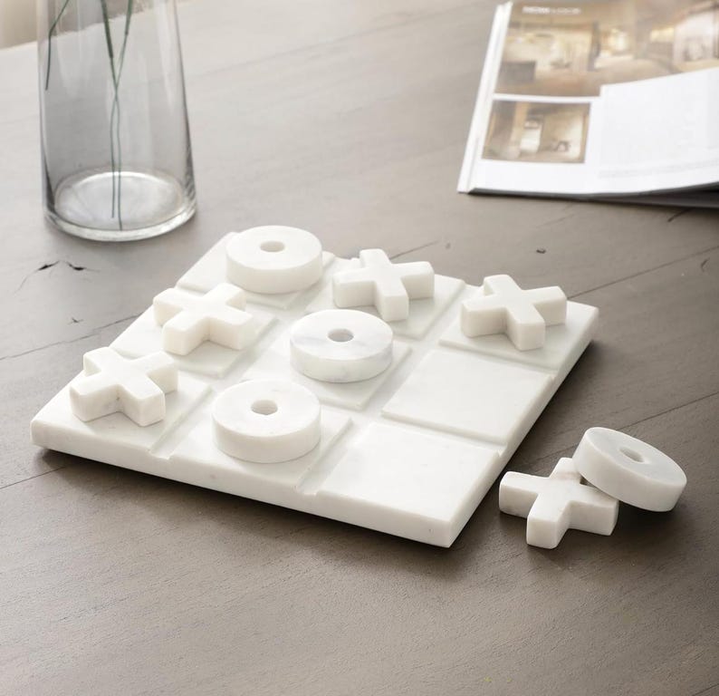 May include: A white marble tic-tac-toe game set. The game board features a grid with 'X' and 'O' game pieces. The set includes a board and individual game pieces. The game is made of white marble.