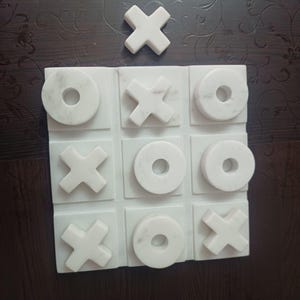 May include: A white marble tic-tac-toe game set. The game board features a grid of nine squares, with "X" and "O" game pieces. The set includes a single "X" piece placed above the board. The marble has a natural veining pattern.