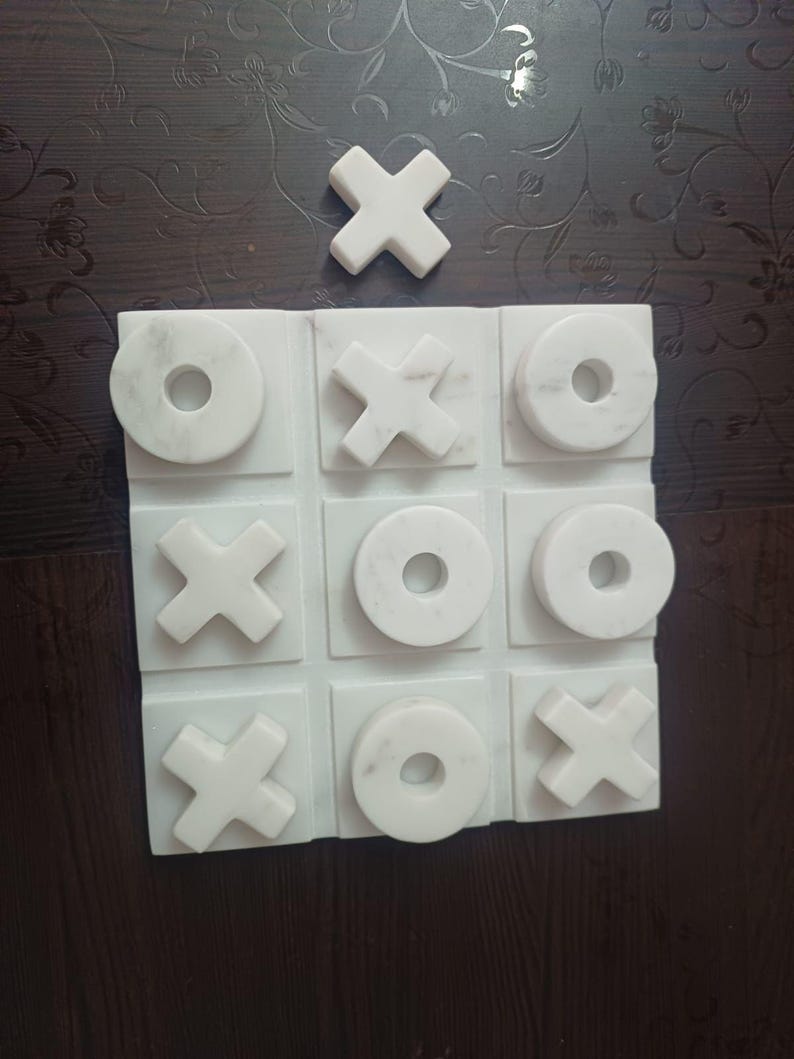 May include: A white marble tic-tac-toe game set. The game board features a grid of nine squares, with "X" and "O" game pieces. The set includes a single "X" piece placed above the board. The marble has a natural veining pattern.