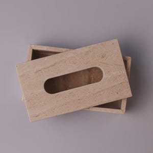May include: A rectangular tissue box cover crafted from beige travertine stone with a natural porous texture. The cover features a centered, elongated oval opening for dispensing tissues, showcasing the unique veining and earthy tones of the stone.