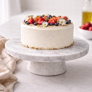 Puede incluir: Un pastel redondo glaseado blanco decorado con fresas frescas, arándanos y pequeñas flores blancas. El pastel se encuentra sobre un soporte para pasteles de mármol blanco. El pastel tiene un borde desmenuzable y está sobre una superficie de mármol blanco.