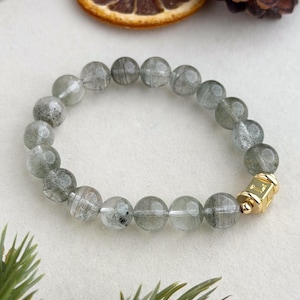 Puede incluir: Un brazalete de cuentas translúcidas de color gris verdoso, con un cierre dorado. Las cuentas tienen aproximadamente 1 cm de diámetro. El brazalete se exhibe sobre una superficie blanca con elementos decorativos.