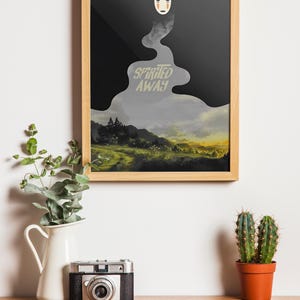 K&ouml;nnte beinhalten: Gerahmter Kunstdruck mit einer Szene aus "Chihiros Reise ins Zauberland". Das Kunstwerk hat einen schwarzen und grauen Hintergrund mit dem Filmtitel in einer stilisierten Schriftart. Oben befindet sich eine wei&szlig;e Maske. Eine Vintage-Kamera, ein wei&szlig;er Krug mit Gr&uuml;n und ein Kaktus stehen auf einer Holzoberfl&auml;che.