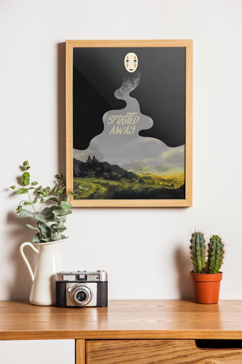 K&ouml;nnte beinhalten: Gerahmter Kunstdruck mit einer Szene aus "Chihiros Reise ins Zauberland". Das Kunstwerk hat einen schwarzen und grauen Hintergrund mit dem Filmtitel in einer stilisierten Schriftart. Oben befindet sich eine wei&szlig;e Maske. Eine Vintage-Kamera, ein wei&szlig;er Krug mit Gr&uuml;n und ein Kaktus stehen auf einer Holzoberfl&auml;che.