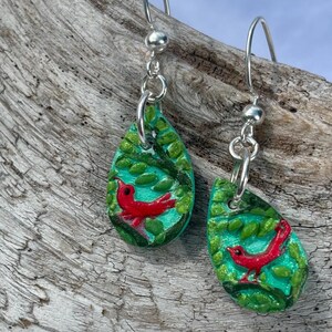 May include: Teardrop-shaped earrings with a red cardinal bird design. The earrings feature a turquoise background with green leaves and silver-colored ear wires. The bird is painted in red, and the leaves are green.