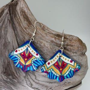 May include: A pair of handmade, blue, red, yellow, and white painted earrings. The earrings have a fan-like shape with intricate patterns and silver-colored ear wires. The earrings are displayed on a piece of driftwood.