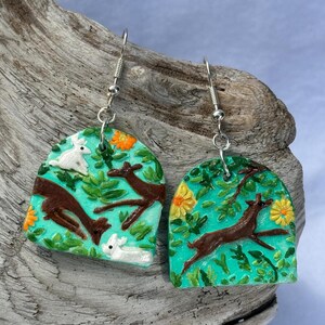 May include: A pair of handcrafted earrings featuring a nature-inspired design. Each earring showcases a teal background with brown deer, white rabbits, green leaves, and orange and yellow flowers. The earrings have silver-colored hooks.