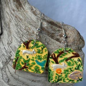 May include: Handmade earrings featuring a whimsical forest scene. Each earring has a yellow background with a white rabbit, a blue bird, and green foliage. The earrings are suspended from silver-colored fish hook ear wires.