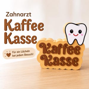 May include: A tan, cloud-shaped sign with the brown text 'Kaffee Kasse' and a white, smiling tooth icon with black eyes and pink cheeks. A separate tan label features a brown heart icon and the text 'Für ein Lächeln bei jedem Besuch!'