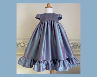 Peut inclure: Robe de cérémonie bleu et violet irisé, présentée sur un mannequin. Le corsage est en tissu gris texturé, avec des manches courtes. La jupe est en matière brillante et fluide avec un ourlet à volants. La robe est pour enfant.