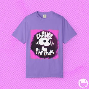 May include: A lilac-colored t-shirt featuring a graphic design. The design includes the words "Change Threads" in white, set against a dark, textured background with a pink border. The shirt has short sleeves and a crew neck.