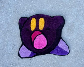 May include: A handmade, plush rug shaped like a cartoon character. It has a dark purple body, yellow eyes, a pink mouth, and purple feet. The rug is on a grey concrete surface.