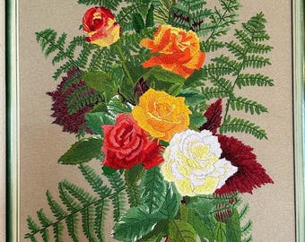 Op de afbeelding: Geborduurde bloemenkunst met een boeket rozen in tinten rood, geel en wit, omringd door groene varens en bladeren. Het kunstwerk is ingelijst in een groene houten lijst.