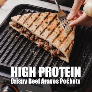 Può includere: Un sandwich triangolare grigliato, ripieno di carne macinata, appoggiato su una griglia nera. Il panino è etichettato "HIGH PROTEIN Crispy Beef Arayes Pockets". Una forchetta viene utilizzata per tagliare il panino. Il cibo sembra appena fatto.