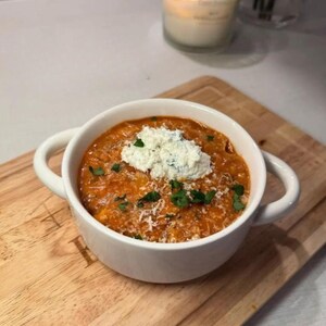 May include: A white bowl filled with a vibrant orange-red soup, topped with a dollop of white cheese and sprinkled with green herbs. The bowl has two handles and sits on a wooden cutting board. A lit candle is in the background.
