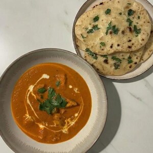 Könnte beinhalten: Eine Schüssel mit orangefarbenem Curry, verziert mit Sahne und grünen Kräutern, steht neben einem Teller mit Naan-Brot, das mit Kräutern bestreut ist. Das Essen wird auf einer weißen und grauen Marmoroberfläche präsentiert.