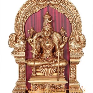 Può includere: Una statua dorata di una divinità, seduta all'interno di un arco ornato. La statua è molto dettagliata, con intagli e abbellimenti intricati. Lo sfondo presenta una tenda rossa e la base è di colore marrone scuro. È un oggetto religioso.
