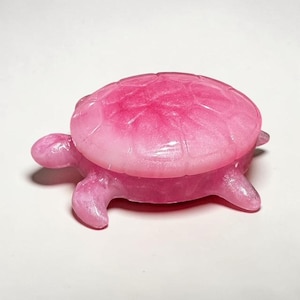 May include: A small, decorative turtle figurine crafted from a translucent, shimmering pink material. The turtle features a smooth, rounded shell with visible segmented patterns and four extended flippers, all in a consistent bright pink hue.