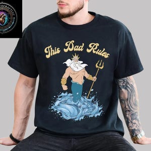 May include: Black t-shirt with the text "This Dad Rules" in gold lettering. The shirt features a cartoon illustration of a merman holding a trident, emerging from blue water. The Voidpulsecraftllc logo is in the upper left corner.