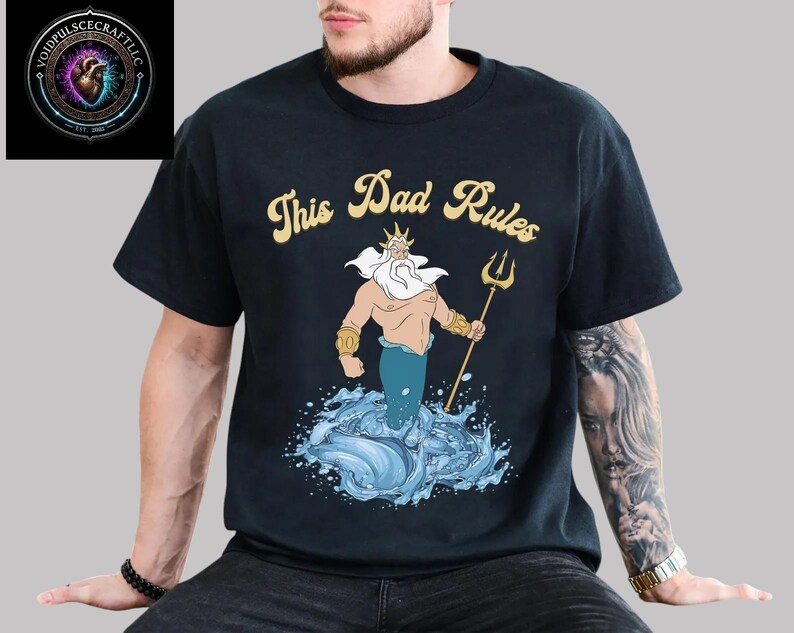 May include: Black t-shirt with the text "This Dad Rules" in gold lettering. The shirt features a cartoon illustration of a merman holding a trident, emerging from blue water. The Voidpulsecraftllc logo is in the upper left corner.