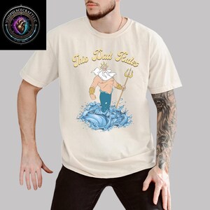 May include: Cream-colored t-shirt featuring the phrase "This Dad Rules" above a cartoon illustration of a trident-wielding sea god rising from blue waves. The design includes gold, blue, and tan colors. The shirt is short-sleeved.