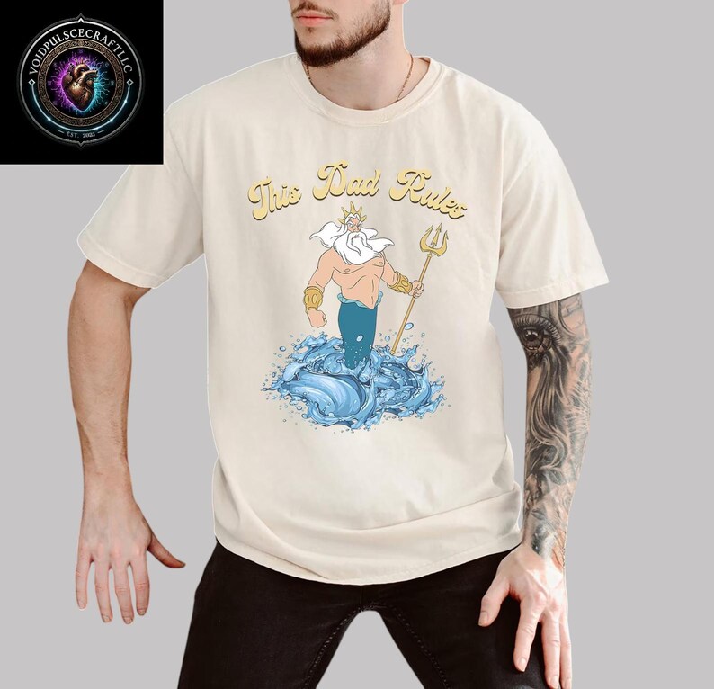 May include: Cream-colored t-shirt featuring the phrase "This Dad Rules" above a cartoon illustration of a trident-wielding sea god rising from blue waves. The design includes gold, blue, and tan colors. The shirt is short-sleeved.
