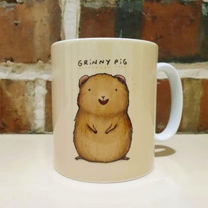 May include: A beige ceramic mug with a brown and white illustration of a smiling guinea pig. The mug has the text "Grinny Pig" printed on it.