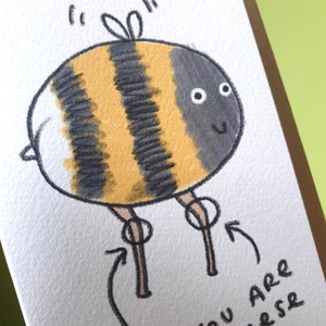 Puede incluir: Una ilustraci&oacute;n dibujada a mano de una abeja de dibujos animados con rayas negras y amarillas, alas blancas y patas marrones. La abeja sonr&iacute;e y tiene dos ojos grandes y redondos. El texto "You are a bee-r" est&aacute; escrito debajo de la abeja.