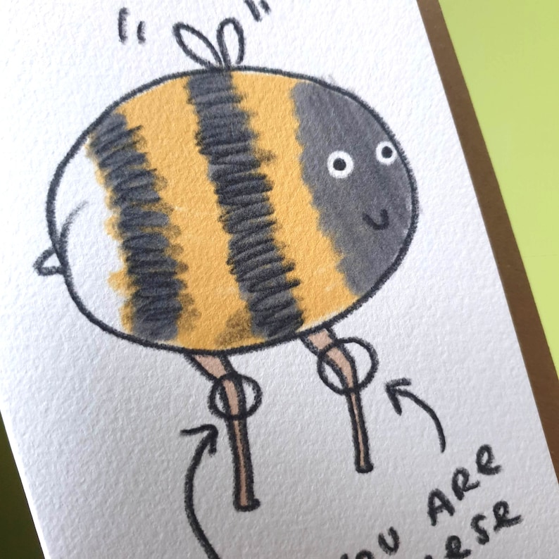 Puede incluir: Una ilustraci&oacute;n dibujada a mano de una abeja de dibujos animados con rayas negras y amarillas, alas blancas y patas marrones. La abeja sonr&iacute;e y tiene dos ojos grandes y redondos. El texto "You are a bee-r" est&aacute; escrito debajo de la abeja.
