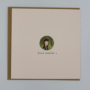 May include: A white card with a brown border. The card has a circular illustration of a woman with brown hair in a forest setting. The text "Sophie Corrigan x" is written in black ink below the illustration.