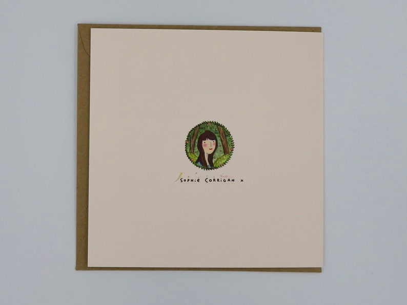 May include: A white card with a brown border. The card has a circular illustration of a woman with brown hair in a forest setting. The text "Sophie Corrigan x" is written in black ink below the illustration.
