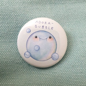 Puede incluir: Un botón blanco con un personaje de burbuja azul con una cara sonriente y el texto "ADORA-BUBBLE" en él.