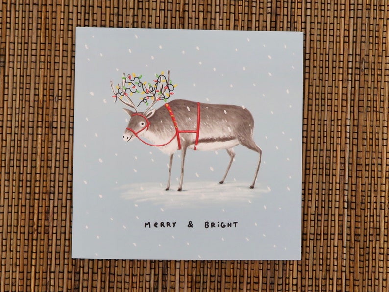 K&ouml;nnte beinhalten: Weihnachtskarte mit einem Rentier mit Lichterketten auf dem Geweih, rotem Geschirr und einer Schneelandschaft. Die Worte "MERRY & BRIGHT" stehen unten auf der Karte.