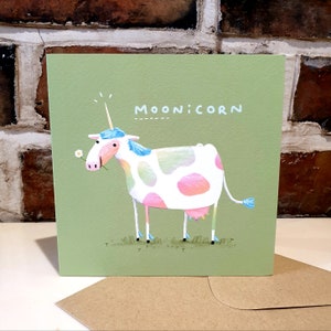 May include: A green card with a cartoon illustration of a pink and white cow with a blue unicorn horn and a blue tail. The cow is holding a white flower in its mouth. The text "MOONICORN" is written in white on the card.