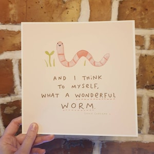Könnte beinhalten: Ein rosa-weißer Wurm mit einem fröhlichen Gesicht ist auf einem hellrosa Hintergrund gezeichnet. Der Text lautet "Und ich denke mir, was für ein wundervoller Wurm." Der Text ist in Braun und der Name der Künstlerin, "Sophie Corrigan", steht in kleinerer Schrift unter dem Text.