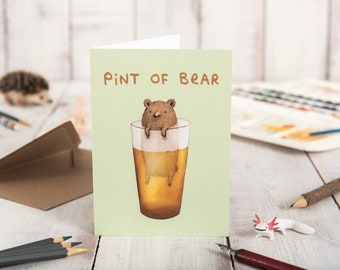 May include: A greeting card with a whimsical illustration. The card features a bear inside a pint glass of beer, with the text "PINT OF BEAR" above. The card has a light green background and is set on a white wooden surface.