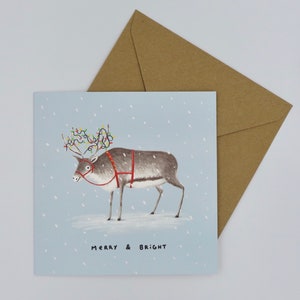 K&ouml;nnte beinhalten: Weihnachtskarte mit einem Rentier, das bunte Lichterketten auf dem Geweih und einen roten Geschirr tr&auml;gt. Auf hellblauem Hintergrund steht &bdquo;MERRY & BRIGHT&ldquo;. Ein brauner Umschlag liegt daneben.