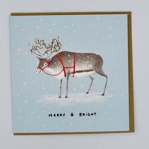 K&ouml;nnte beinhalten: Weihnachtskarte mit einem Rentier, das mit Lichterketten auf seinem Geweih und einem roten Geschirr geschm&uuml;ckt ist. Das Rentier steht in einer verschneiten Szene mit dem Schriftzug "MERRY & BRIGHT" darunter. Die Karte hat einen hellblauen Hintergrund und einen braunen Rand.