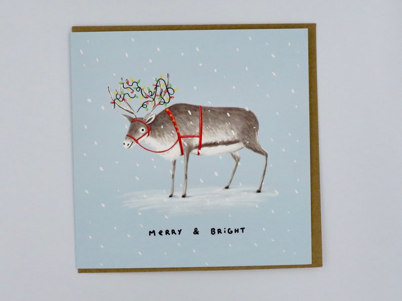 K&ouml;nnte beinhalten: Weihnachtskarte mit einem Rentier, das mit Lichterketten auf seinem Geweih und einem roten Geschirr geschm&uuml;ckt ist. Das Rentier steht in einer verschneiten Szene mit dem Schriftzug "MERRY & BRIGHT" darunter. Die Karte hat einen hellblauen Hintergrund und einen braunen Rand.