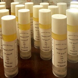 May include: A collection of lip balms in clear tubes with white caps and yellow bands. Each tube has a label with the text "Monoi Butter" and "Nassie's Hope Lip Balm" for soft and smooth lips. Each tube contains .15 ounces.