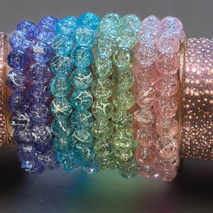 May include: A set of four stretch bracelets featuring round crackle glass beads in a gradient of colors including purple, blue, green, and pink. Each bead displays a distinct internal webbed texture with white veining throughout the surface.