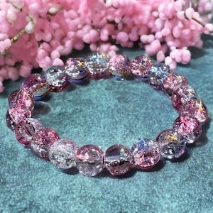 May include: A beaded bracelet with clear, pink, and purple beads. The bracelet is circular and sits on a teal surface. Pink flowers are in the background, adding a soft contrast to the jewelry.