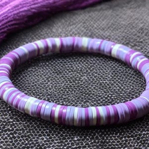 May include: A purple bracelet with a mix of light purple, white, and dark purple disc-shaped beads. The bracelet is circular and sits on a textured gray surface. A purple fabric is visible in the background.