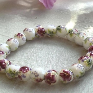 Pode incluir: Uma pulseira de contas brancas com desenhos florais em tons de roxo, verde e vermelho. A pulseira &eacute; feita de contas redondas, cada uma adornada com um padr&atilde;o floral diferente. &Eacute; uma joia.