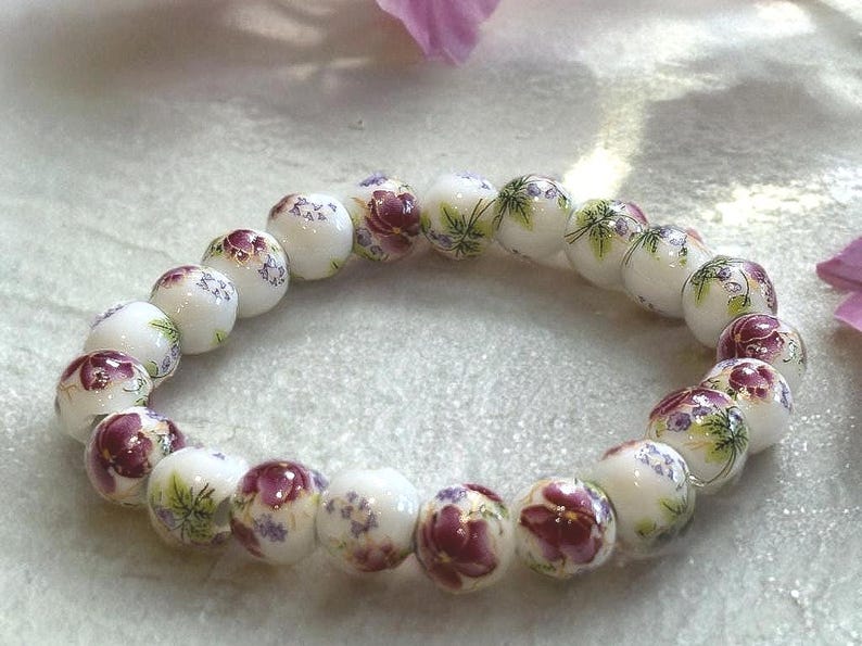 Pode incluir: Uma pulseira de contas brancas com desenhos florais em tons de roxo, verde e vermelho. A pulseira &eacute; feita de contas redondas, cada uma adornada com um padr&atilde;o floral diferente. &Eacute; uma joia.