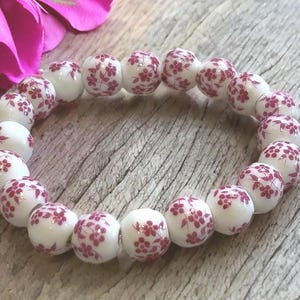 May include: A white beaded bracelet with a floral pattern in shades of red. The bracelet is made of round beads and is displayed on a wooden surface, with a pink flower in the background.