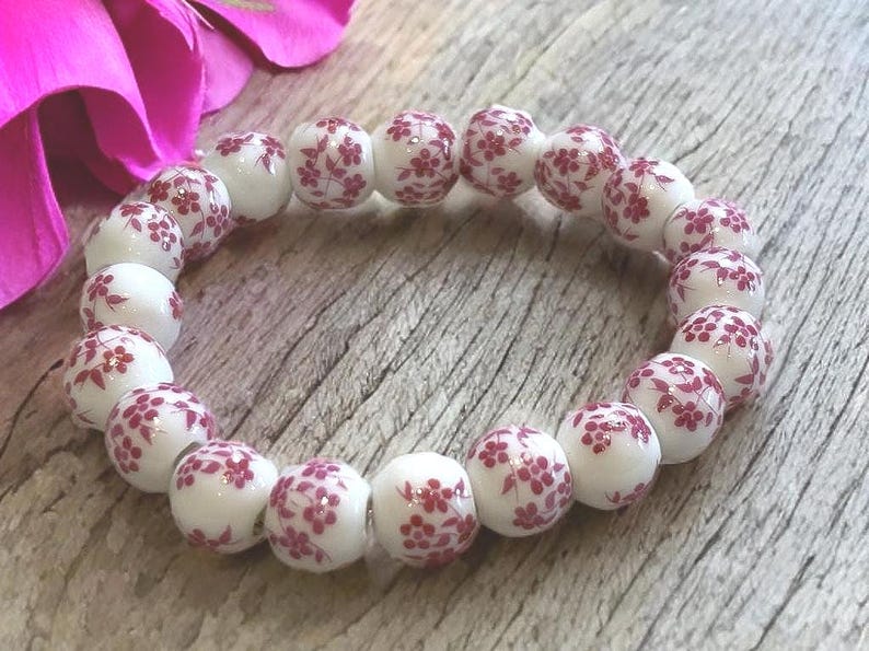 Pode incluir: Uma pulseira de contas brancas com um padr&atilde;o floral vermelho. A pulseira, composta por contas redondas, &eacute; apresentada numa superf&iacute;cie de madeira. Uma flor rosa &eacute; vis&iacute;vel no fundo.