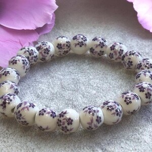 Pode incluir: Uma pulseira de contas de cer&acirc;mica branca com um padr&atilde;o floral roxo. As contas redondas s&atilde;o enfiadas, criando uma forma circular. A pulseira &eacute; exibida em uma superf&iacute;cie cinza, com flores roxas no fundo.