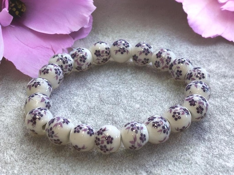 Pode incluir: Uma pulseira de contas de cer&acirc;mica branca com um padr&atilde;o floral roxo. As contas redondas s&atilde;o enfiadas, criando uma forma circular. A pulseira &eacute; exibida em uma superf&iacute;cie cinza, com flores roxas no fundo.