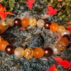 May include: A stretch bracelet composed of alternating round beads in shades of dark brown, bright orange, and translucent beige, interspersed with gold-toned glitter-textured spherical beads arranged in a repeating circular pattern.